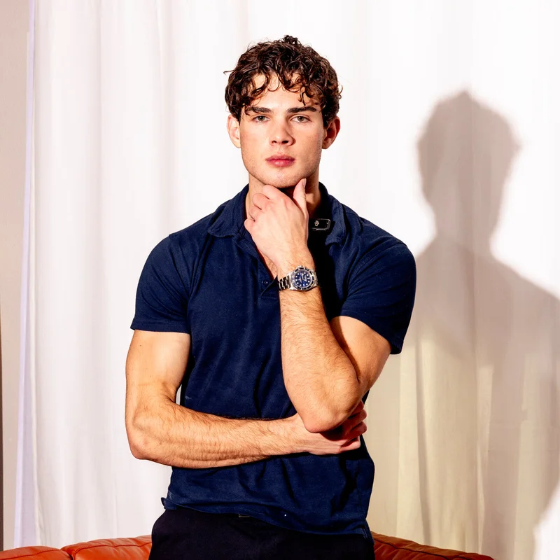 Le streamer Clavicular en polo bleu marine avec une montre au poignet, assis sur un canap&eacute; marron devant un fond blanc, connu pour son contenu lookmaxxing sur Kick.