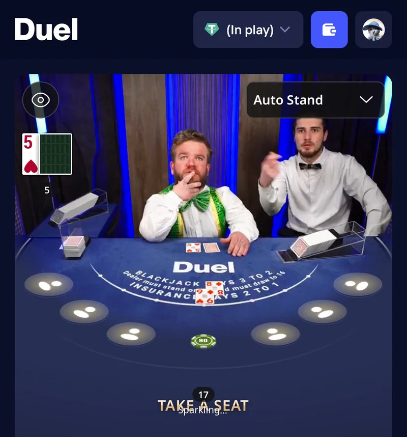Table de blackjack live sur Duel Casino avec deux croupiers en studio, l'un en chemise blanche avec un n&oelig;ud papillon vert et jaune, l'autre en tenue classique, pendant une vraie partie film&eacute;e en direct